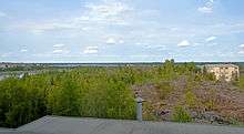 A largely level landscape of short evergreen trees and exposed grayish bedrock. In the foreground, at bottom, is a roof with a shiny metal circular pipe. At rear left is a lake; there are some buildings and a Canadian flag at the rear right