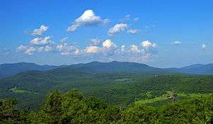 A range of mountains, mostly rounded and covered with green woods, seen past fields in the distance. Above them is a blue sky filled with little fluffy clouds.