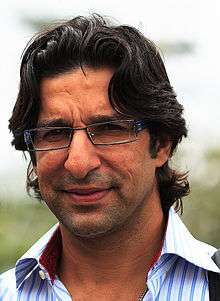 A headshot of an Asian man facing forwards.  He has shoulder-length, dark, wavy hair and is wearing a blue and white-striped shirt.  He is also wearing near-rectangular-framed spectacles.