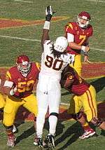 An American football player in a white, gold,and brown uniform reaches to block a pass by another player. He is being blocked by two other players in red and yellow uniforms.