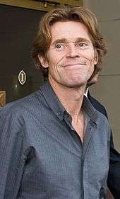 A portrait of Willem Dafoe wearing a blue shirt.