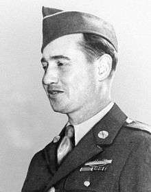 Head and shoulders of a white man wearing a garrison cap tilted over his right ear and a military jacket with a badge and two ribbon bars on the left breast.