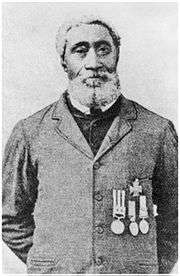 A black-and-white photograph of a man with a beard and short hair looking at the viewer and wearing a button-up jacket displaying four medals