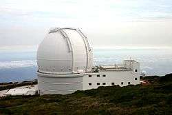 View of large all white building with the biggest part being a spherical shaped section placed atop a rectangular prism. The building sits atop a mountain with clouds showing in the distance above and below the height of the building.