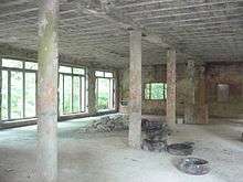 An empty room in an abandoned building with no finishes, cement structural systems visible, support columns in the middle, empty openings along a left brick wall through which a forest can be seen, and cement blocks and tires piled on the floor